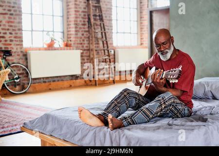 Uomo con occhiali che suonano la chitarra a letto a casa Foto Stock