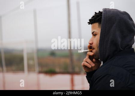 Giovane uomo che indossa una camicia con cappuccio fumando una sigaretta Foto Stock