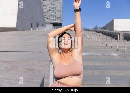 Donna sorridente e curvilinea che indossa abbigliamento sportivo in piedi con gli occhi chiusi sulle scale Foto Stock
