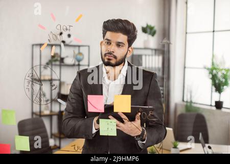 Dipendente dell'azienda che si trova in un ufficio moderno e che guarda il diagramma finanziario su un pannello di vetro. Lavoratore indiano maschile in tuta formale che si trova a capire le strategie di investimento con il tablet digitale in mano. Foto Stock