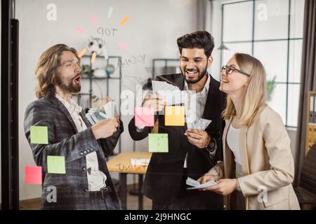 Gioiosi partner multietnici che detengono denaro guadagnato in contanti mentre si trova vicino a un pannello di vetro con note appiccicose. Felice gente ricca di affari nelle cause che celebra la crescita di reddito e successo finanziario. Foto Stock