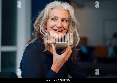 Donna anziana felice che invia messaggi vocali tramite smartphone in ufficio Foto Stock