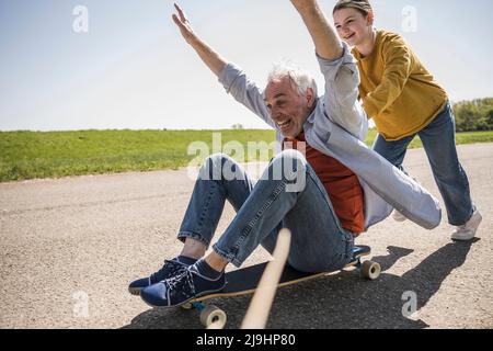 Ragazza giocosa che spinge il nonno allegro seduto con le braccia sollevate sullo skateboard Foto Stock