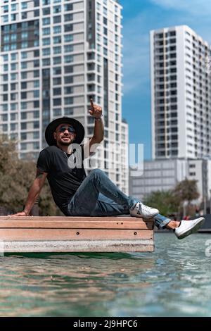 il giovane uomo gode di una giornata di sole e punta a qualcosa nel mezzo di un fiume nel mezzo della città circondato da edifici. Foto Stock