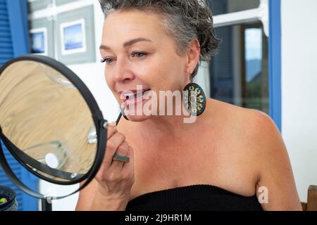 Donna matura applicazione rossetto seduta di fronte allo specchio a casa Foto Stock