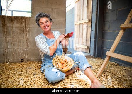 Donna contadina felice matura con gallina e ciotola d'uovo seduta in polpaccio Foto Stock