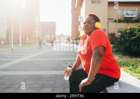 Donna seduta con bottiglia d'acqua che ascolta la musica tramite cuffie wireless Foto Stock