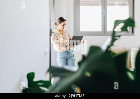 Giovane donna che usa un computer portatile in piedi in salotto a casa Foto Stock