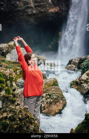 Felice giovane donna con le braccia sollevate in piedi di fronte alla cascata Foto Stock