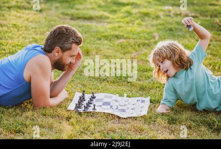 Padre e figlio che giocano a scacchi sdraiati sull'erba al parco del prato. Festa dei padri, famiglia d'amore, genitori, concetto d'infanzia. Foto Stock