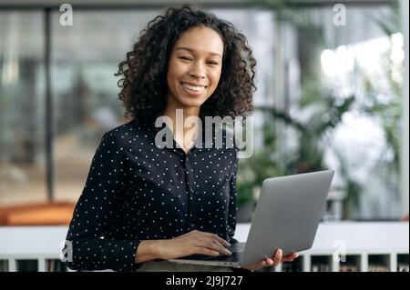 Ritratto di successo ragazza ricci con pelle scura, analista finanziario, auditor, specialista IT, si alza in ufficio, in abiti da lavoro, tiene un laptop aperto nelle sue mani, guarda la fotocamera, sorride Foto Stock