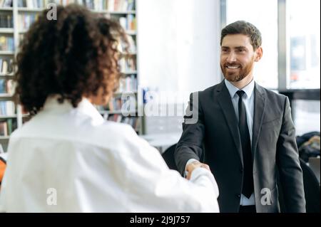 Cooperazione commerciale. Fare un affare, scuotendo le mani. L'uomo d'affari caucasico, capo dell'azienda, scuote le mani con la sua partner d'affari afro-americana, conferma accordi, sorridono l'un l'altro Foto Stock