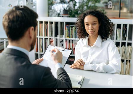 La ragazza afroamericana intenzionale, in camicia bianca, è intervistando per un lavoro, si presenta, il responsabile di assunzione esamina attentamente il riassunto, fa domande, analizza, fa scelte, prende appunti Foto Stock