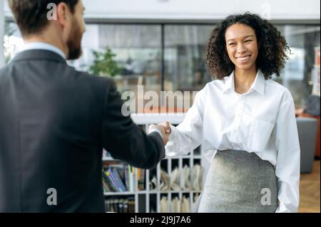 Fare un accordo, scuotendo le mani, conferma gli accordi. Donna d'affari di successo afro-americana, responsabile delle vendite, scuote le mani con il collega maschio o il cliente in ufficio moderno, sorridendo a vicenda Foto Stock