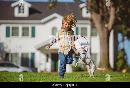 Cane con guinzaglio che corre con gestore. Ragazzino sullo sfondo di un prato verde nel cortile in una giornata di sole estiva. Piccolo cucciolo che insegue il bambino. Foto Stock