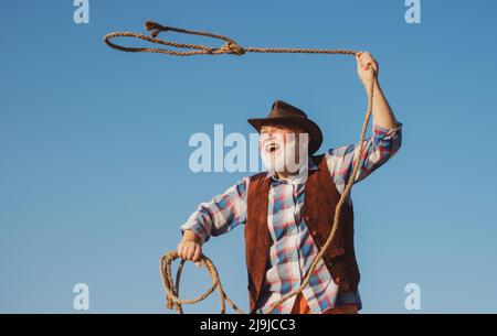 Vecchio cowboy selvaggio West con corda. Uomo occidentale bearded gettando lazo con giacca marrone e cappello che cattura cavallo o mucca. Foto Stock