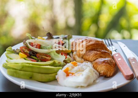 Sandwich con croissant con uova in camicia, pomodoro e avocado sul piatto in caffetteria, da vicino Foto Stock