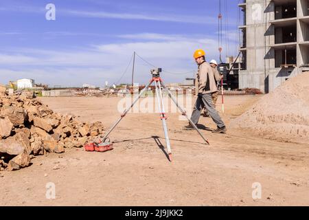 Kemerovo, Russia - Jule 02 2021. La livella ottica è installata in cantiere vicino a un mucchio di pietra grande, il lavoratore in caschi passa vicino, lavori di topografia Foto Stock