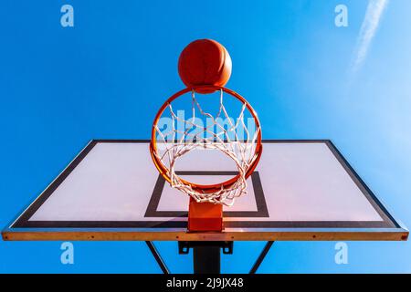 Palla di basket arancione che cade nel cerchio. Urban gioco giovanile. Concetto di successo, punteggio e vincita. Vista ad angolo basso. Foto Stock