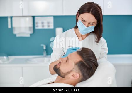 Cosmetologo con maschera fa iniezione di medicina al cliente durante la terapia anti-invecchiamento meso in salone Foto Stock