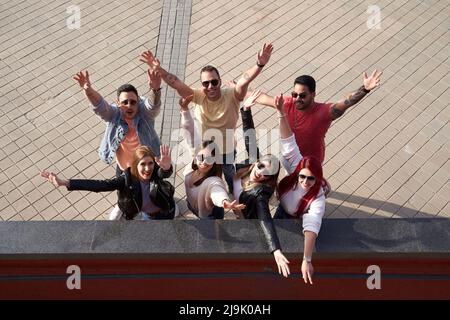 Da sopra gruppo di giovani allegri amici multirazziali maschili e femminili in abiti casual che alzano le braccia e sorridono felicemente mentre si alza in piedi su strada lastricata il giorno di sole Foto Stock