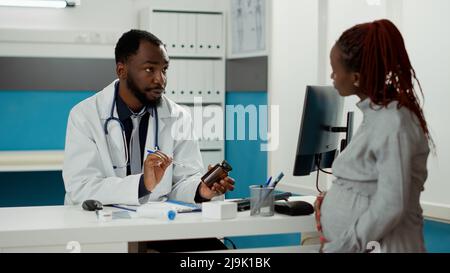 Medico generico che spiega il trattamento di prescrizione in bottiglia a donna che si aspetta il bambino. Specialista in gravidanza utilizzando farmaci pillole per aiutare la paziente in gravidanza con parto in gabinetto. Foto Stock