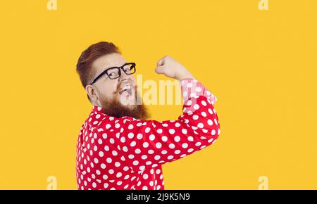 Divertente ed elegante uomo barbato con sorriso sui visi che mostra bicipiti su sfondo giallo. Foto Stock