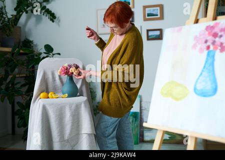 Donna anziana in abbigliamento casual che corregge i fiori in vaso mentre dipinge la vita a classe Foto Stock