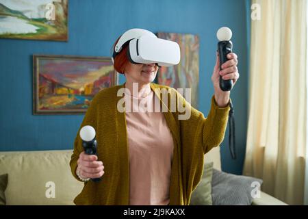 Donna matura in occhiali di protezione che tiene i joystick nelle sue mani e gesturing durante il gioco di realtà virtuale a casa Foto Stock