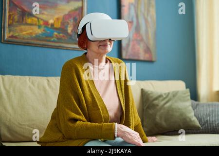 Donna matura seduta sul divano in occhiali VR e guardando qualcosa di virtualità Foto Stock