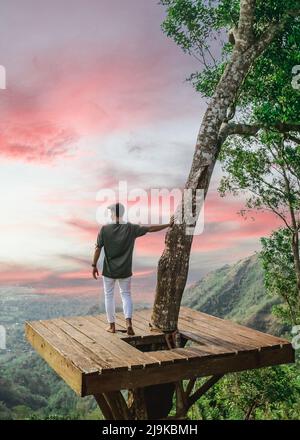 Giovane uomo in piedi su una piattaforma di legno treehouse che tiene su un albero che domina una valle durante il tramonto a Bali Indonesia Foto Stock