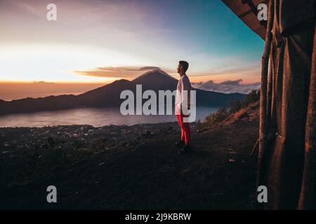 Giovane turista maschile in piedi sulla cima del vulcano Batur durante l'alba in pantaloni rossi e felpa bianca con cappuccio che si affaccia sul lago Foto Stock