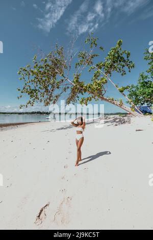 Giovane ragazza asiatica deserta su una spiaggia tropicale di sabbia bianca in un costume da bagno bikini bianco con una tenda blu sull'isola e albero che cade sopra di lei Foto Stock