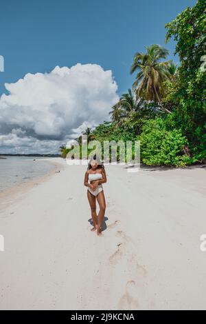 Giovane ragazza asiatica in bikini bianco su una spiaggia di sabbia bianca dell'isola tropicale circondata da palme da cocco in una giornata tropicale di sole Foto Stock