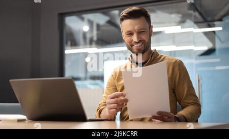 Felice uomo d'affari che legge i documenti di affari che si siedono vicino al laptop in ufficio Foto Stock