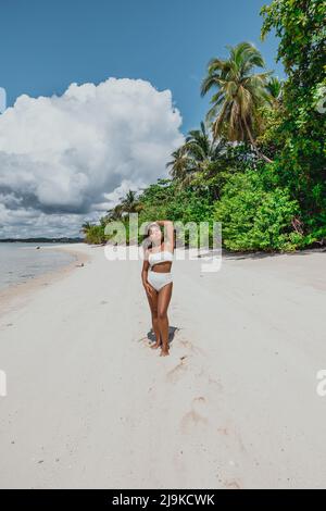 Giovane ragazza asiatica in bikini bianco su una spiaggia di sabbia bianca dell'isola tropicale circondata da palme da cocco in una giornata tropicale di sole Foto Stock