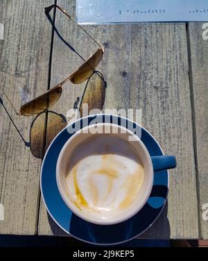 Delizioso e colorato cappuccino cremoso servito in una tazza blu su un tavolo da terrazza con occhiali da sole e menu accanto ad esso Foto Stock