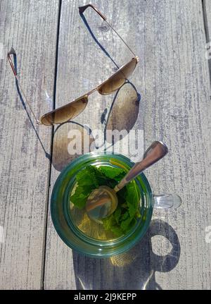 Delizioso tè alla menta verde servito in una tazza calda su un tavolo in legno con occhiali da sole accanto Foto Stock