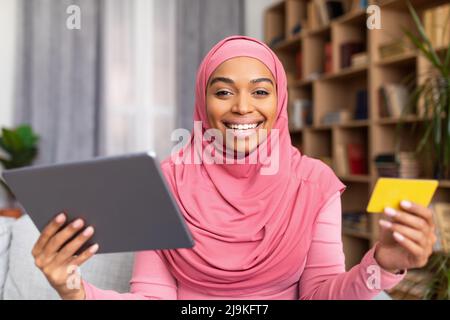 Buona donna musulmana nero shopping da casa, utilizzando tablet digitale e carta di credito, l'acquisto di cose nel negozio web Foto Stock