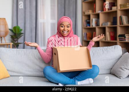 Allegra signora musulmana afroamericana che apre la scatola di cartone, che riceve l'ordine di negozio in linea a casa, seduto sul divano Foto Stock