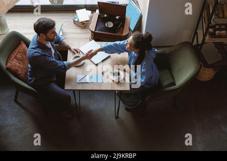 Vista dall'alto su razze miste giovane uomo e donna che si scuotono le mani al tavolo del caffè. Gesto Deal. Foto Stock