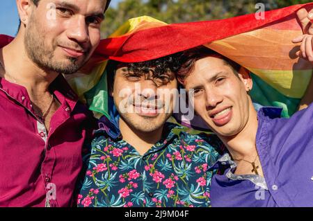 primo piano gay latino caucasico giovani amici maschi, in piedi insieme all'aperto con una bandiera gay orgoglio sopra la testa, guardando la macchina fotografica. Foto Stock