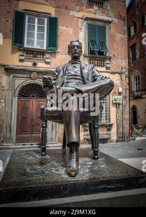 Luuca, Toscana, Italia, 09 maggio 2022 - Statua di Giacomo Puccini all'interno delle mura medievali della città di Lucca, con antico edificio con finestre a ciottoli nella ba Foto Stock