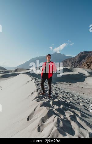 giovane turista maschile in cima a una duna di sabbia con una giacca americana rosso brillante nel deserto freddo di skardu pakistan Foto Stock