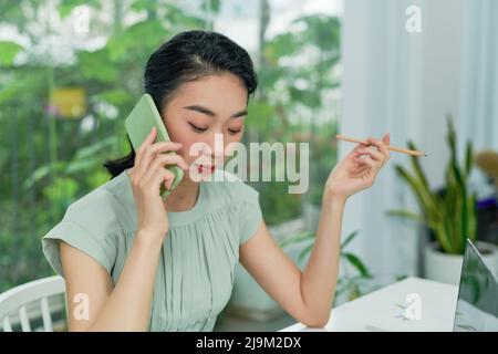 Giovane imprenditrice asiatica che parla al telefono mentre lavora in casa. Foto Stock