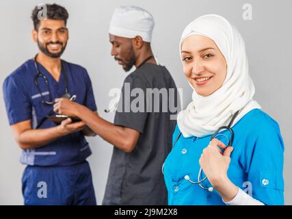 Gruppo di paramedici sorridenti su sfondo grigio. donna medico musulmana con colleghi chirurgo africano e indiano Foto Stock