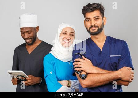 Gruppo di paramedici sorridenti su sfondo grigio. donna medico musulmana con colleghi chirurgo africano e indiano Foto Stock