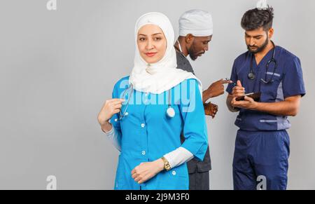 Gruppo di paramedici sorridenti su sfondo grigio. donna medico musulmana con colleghi chirurgo africano e indiano Foto Stock