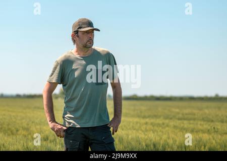 Ritratto di uomo agricolo di mezza età in posa in verde t-shirt e camionista cappello, in piedi in unmipe orzo raccolto in luminoso giorno di primavera soleggiato e. Foto Stock