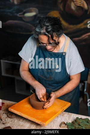 Maturo focalizzato femmina CraftsCeramista donna in grembiule che forma pezzo di argilla pentola in mano in tavolo da officina Foto Stock